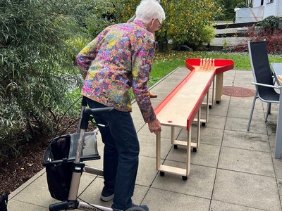 Eine ältere Frau in einer bunten, geblümten Jacke bereitet sich darauf vor, auf einer rot-weißen Holzbahn Kegel zu werfen, begleitet von einem schwarzen Rollator.
