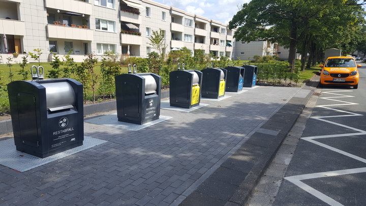 Eine Reihe von sechs schwarz-gelben Recycling-Behältern steht auf einem gemauerten Bürgersteig am Neuenheerser Weg, in dessen Nähe ein gelbes Taxi und ein weißer Lieferwagen parken.