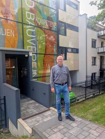 Männliche Person in kariertem Hemd und blauer Jeans steht vor einem modernen Gebäude mit bunter Glasfassade und Schriftzug 'Kompetenz'