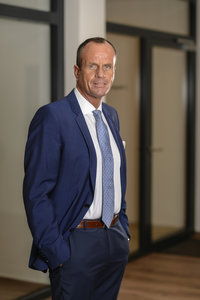 Thorsten Mertens in blauem Anzug, weißem Hemd und blauer Krawatte steht selbstbewusst im Flur der Spar- und Bauverein-Geschäftsstelle, die Hände in den Taschen und ein Lächeln im Gesicht.