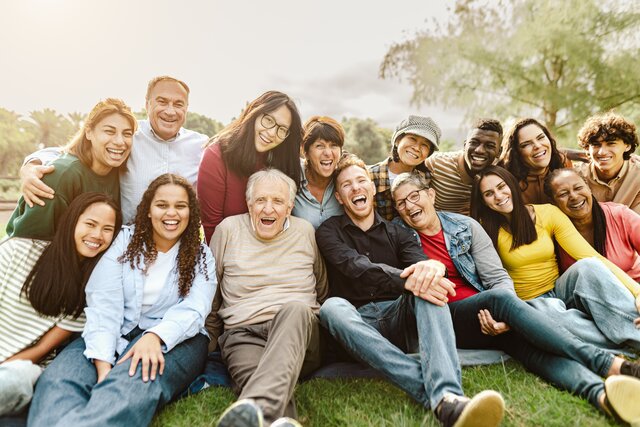 Eine bunte Gruppe von Menschen unterschiedlichen Alters sitzend und lachend auf einer Wiese mit Bäumen im Hintergrund