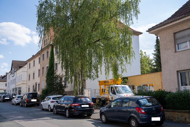 Straße mit parkenden Autos am Fahrbahnrand rechts. Weißes, älteres Mehrfamilienhaus, dass von einer großes flankiert wird.