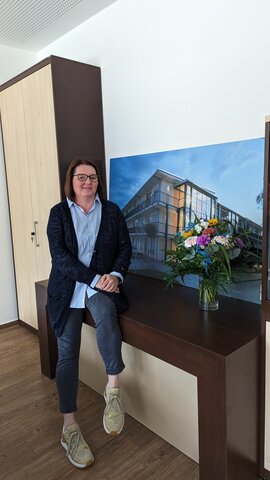 Eine brünette Frau mit Brille sitzt locker auf einer Tischkante neben einer Vase mit bunten Blumen und vor einem großen Bild auf dem ein modernen, hellbeleuchtetes Gebäude zu sehen ist.