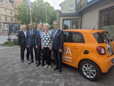 Vier Herren und eine Frau vor einem orangen in Businesskleidung. Im Hintergrund ist ein älteres Gebäude zu sehen.