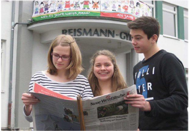 Drei Jugendliche schauen gemeinsam in die Tageszeitung "Neue Westfälische vor dem Eingang ihrer Schule