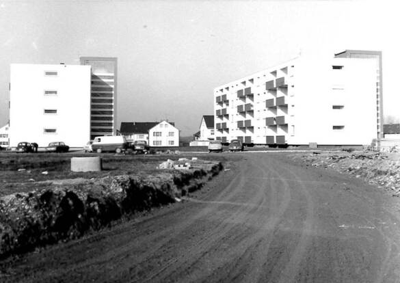 Ein Schwarz-Weiß-Foto zeigt ein Wohngebiet mit weißen Mehrfamilienhäusern, einer unbefestigten Straße und geparkten Autos vor dem Hintergrund eines strahlend blauen Himmels.