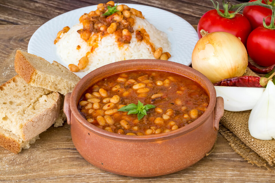 Eine Tonschale mit einem Bohneneintopf, dahinter ein weißer Teller mit Reis und etwas Brot und rechts vom Topf Zwiebeln  und Tomaten