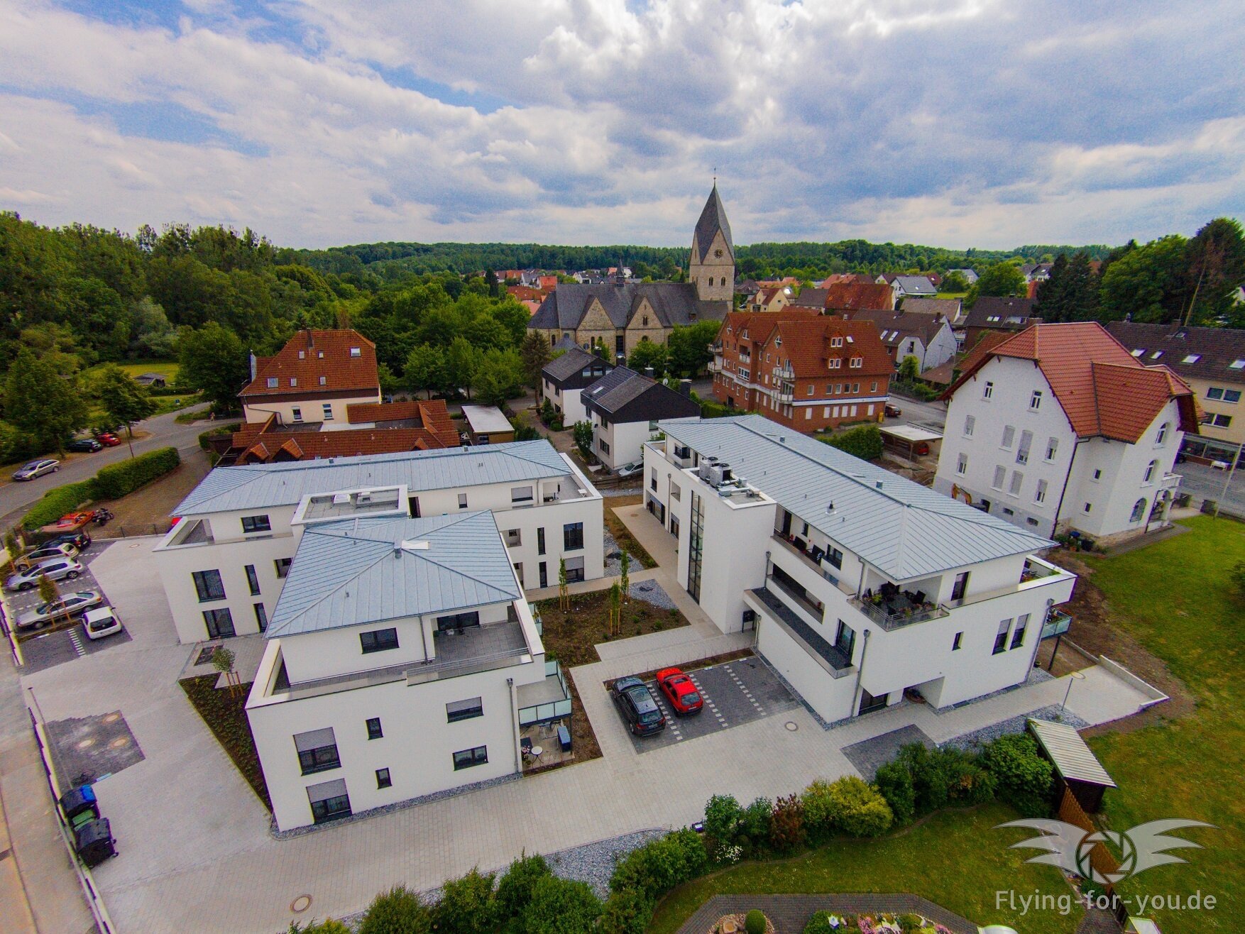 Ein Luftbild von Wewer zeigt eine Mischung aus moderner und traditioneller Architektur mit weißen Gebäuden, roten Dächern und dem Kirchturm, umgeben von üppigem Grün und einem klaren blauen Himmel.