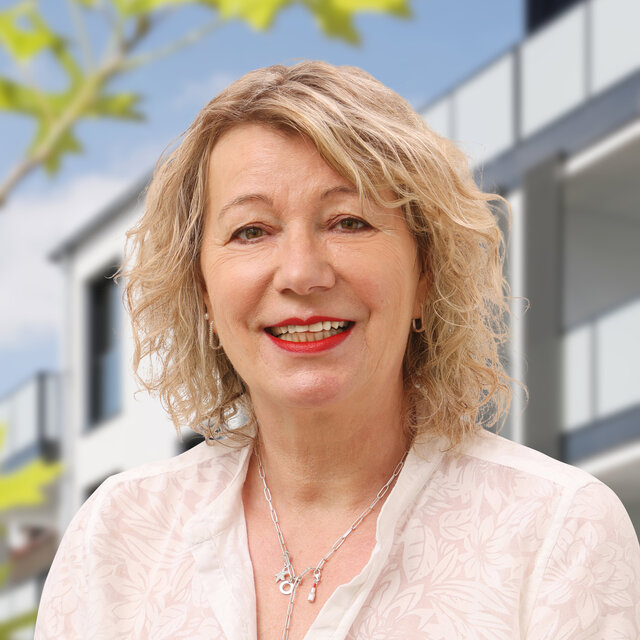 Porträt von Bettina Giese-Walhöfer. Sie schaut lächelnd nach vorn. Sie trägt eine weiße Bluse ohne Kragen und ihre blonden Haare sind halblang und lockig. Im Hintergrund ist die moderne Häuserfassade der Heiersmauer zu erkennen. Grüne Blattelemente sind am linken Rand erkennbar.