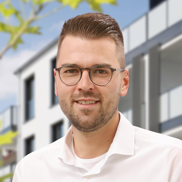 Porträt von Jens Müntefering-Meier. Er schaut lächelnd nach vorn. Er trägt ein weißes Hemd, einen Dreitagebart und einen dunkelblonden Undercut. Im Hintergrund ist die moderne Häuserfassade der Heiersmauer zu erkennen. Grüne Blattelemente sind am linken Rand erkennbar.