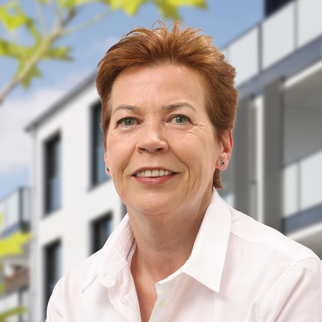 Porträt von Gabi Remmert. Sie schaut lächelnd nach vorn. Sie trägt eine weiße Bluse und ihre brünetten Haare sind kurz geschnitten. Im Hintergrund ist die moderne Häuserfassade der Heiersmauer zu erkennen. Grüne Blattelemente sind am linken Rand erkennbar.