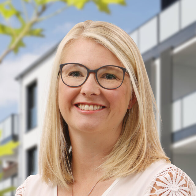 Porträt von Kristin Jacobs. Sie schaut lächelnd nach vorn. Sie trägt eine weiße Bluse mit Spitzenärmeln, eine Brille mit dunklem Gestell und ihre blonden Haare sind mittellang und in der Mitte gescheitelt. Im Hintergrund ist die moderne Häuserfassade der Heiersmauer zu erkennen. Grüne Blattelemente sind am linken Rand erkennbar.