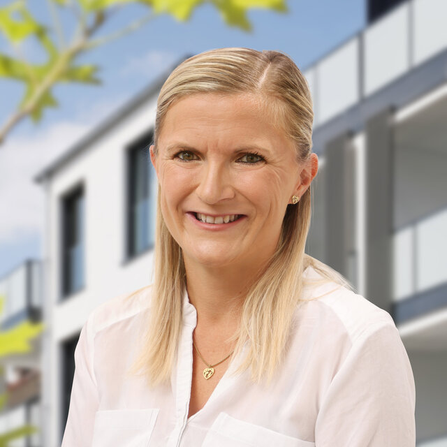 Porträt von Kerstin Dreier. Sie schaut lächelnd nach vorn. Sie trägt eine weiße Bluse ohne Kragen und ihre blonden Haare sind mit einer Spange zurückgenommen. Im Hintergrund ist die moderne Häuserfassade der Heiersmauer zu erkennen. Grüne Blattelemente sind am linken Rand erkennbar.