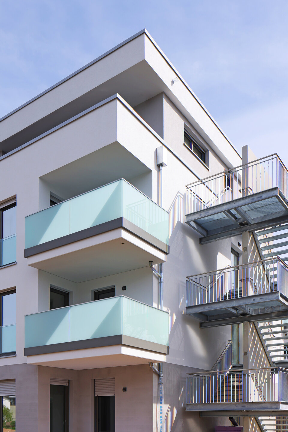 Ein viergeschossiges weißes Gebäude mit Balkonen und einer Glasbrüstung sowie im Erdgeschoss ein kleine Terrasse und rechts davon eine Metalltreppe.