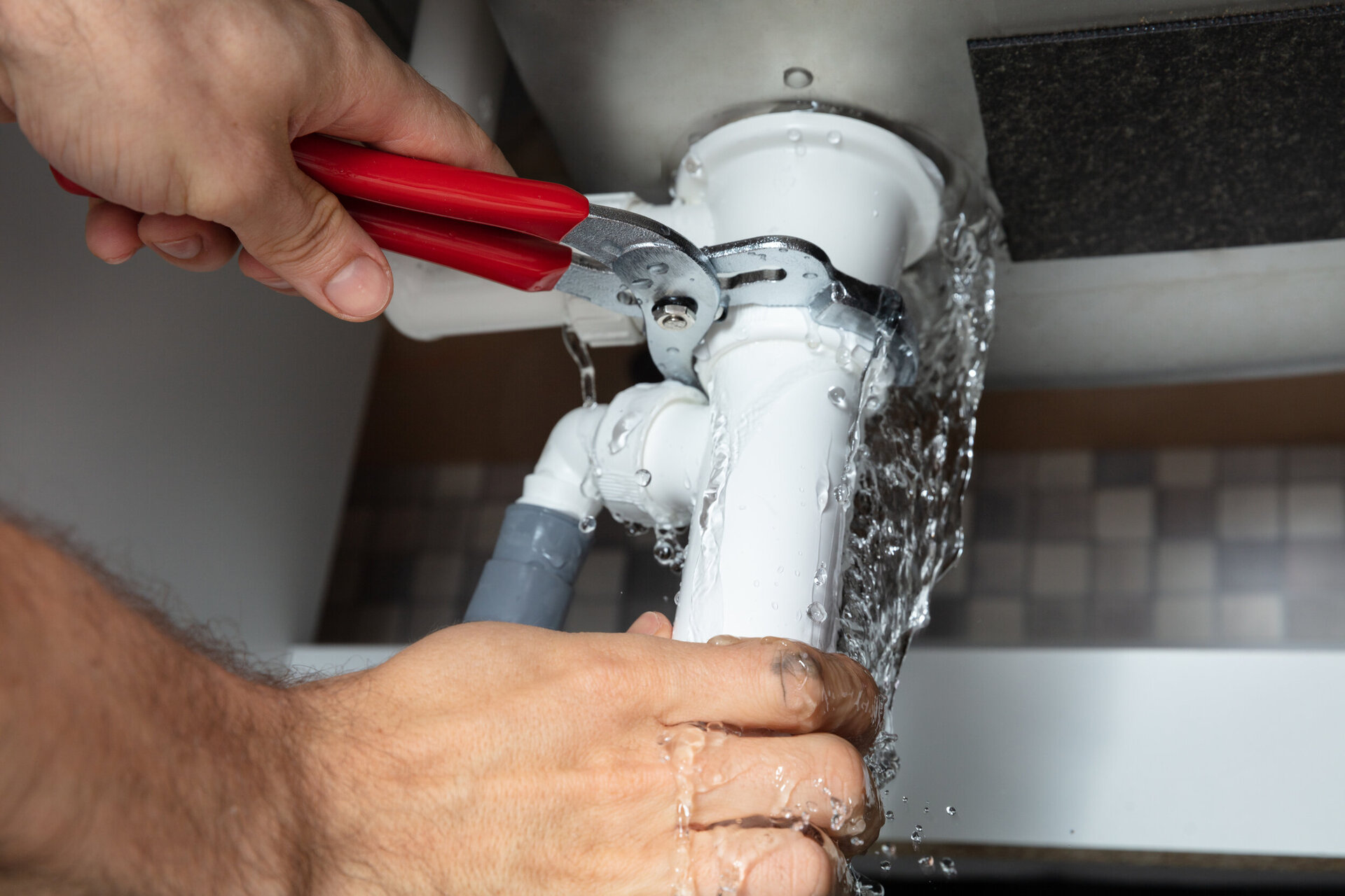 Eine Handwerker benutzt eine Zange, um ein weißes Rohr mit einem silbernen Ventil zu befestigen, das von Wassertropfen auf einer grau gekachelten Wand umgeben ist.