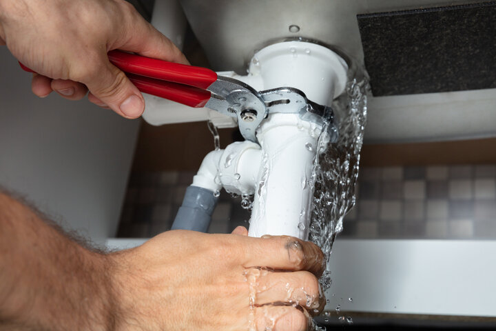 Eine Handwerker benutzt eine Zange, um ein weißes Rohr mit einem silbernen Ventil zu befestigen, das von Wassertropfen auf einer grau gekachelten Wand umgeben ist.