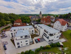 Ein Luftbild von Wewer zeigt eine Mischung aus moderner und traditioneller Architektur mit weißen Gebäuden, roten Dächern und dem Kirchturm, umgeben von üppigem Grün und einem klaren blauen Himmel.