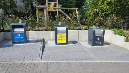 Drei Unterflurcontainer mit den Aufschriften "Papier, Kunststoff, Glas, Metall, Papier, Kunststoff, Glas" stehen in einer Reihe auf einem gepflasterten Platz am Kleinenberger Weg, mit dem Spielplatz und grünen Bäumen im Hintergrund.