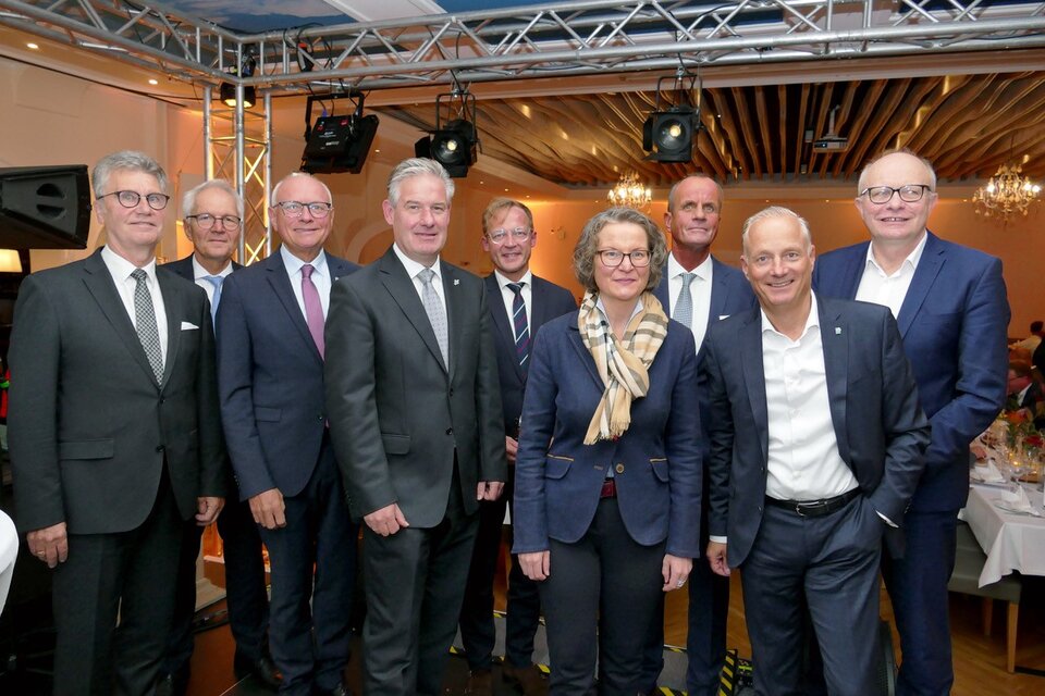 Gruppenfoto mit sieben Herren in Anzügen und einer Frau mit blauem Hosenanzug und braungemusterten Karoschal.