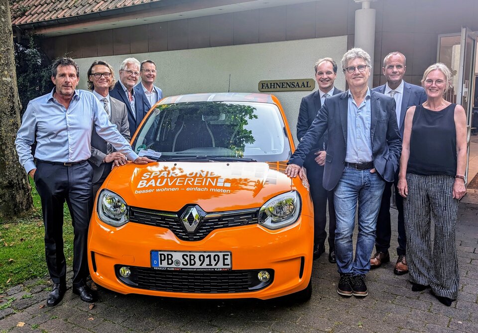 Eine Gruppe von Männern und Frauen flankieren einen orangen Kleinwagen.