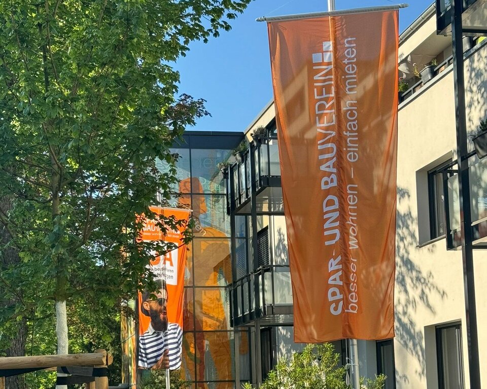 Ein Gebäude, vor dem zwei Fahnenmasten mit orangen Fahnen stehen.
