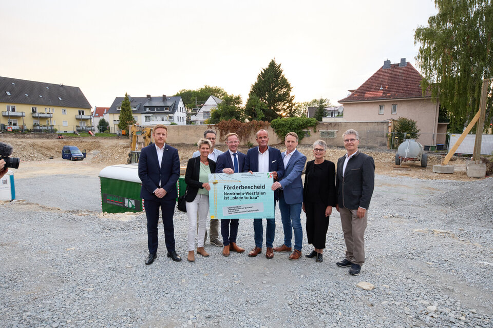 Sieben Personen, zwei Frauen fünf Männer, stehen auf einer Baustelle. Zwei Männer davon halten mittig ein Plakat.