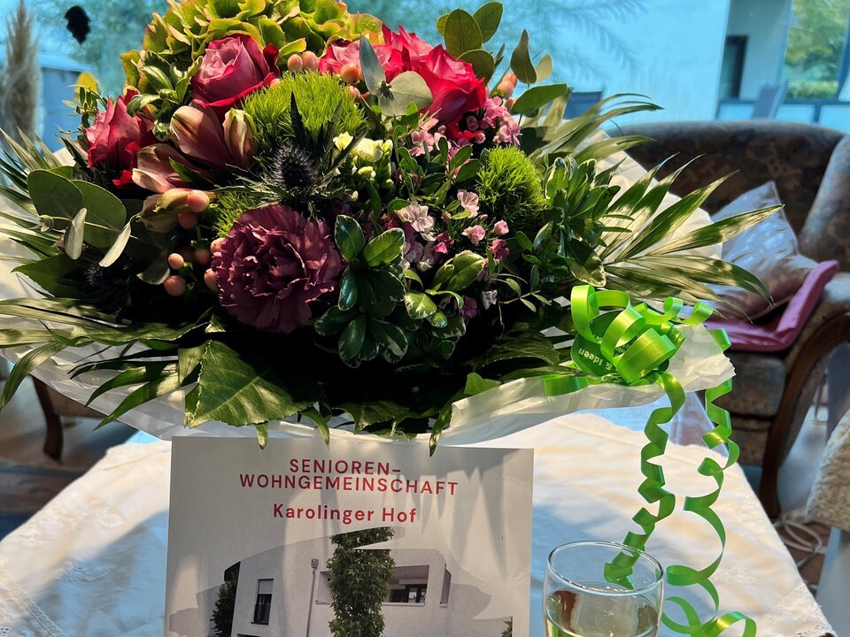 Ein Strauß roter, rosafarbener und lilafarbener Blumen und ein grünes Band liegen auf einem weißen Tischtuch, dazu die Einladung und ein Glas Sekt.