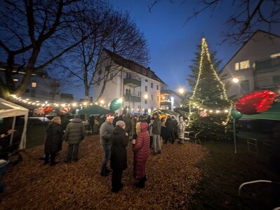 Ein mit weißen Lichtern und roten Ornamenten geschmückter Weihnachtsbaum steht im Innenhof des Quartiers TegelBogen, umgeben von festlich gekleideten Menschen und einem weißen Gebäude mit einem Balkon.