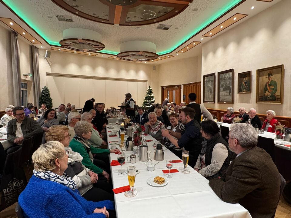Eine Gruppe von langjährigen Mietern des Spar- und Bauvereins sitzt an einem langen Tisch im Festsaal des Schützenhofes Paderborn, unterhält sich und genießt das Essen. Im Hintergrund steht ein großer, festlich geschmückter Weihnachtsbaum.