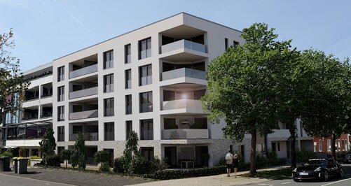 Ein modernes, weißes Mehrfamilienhaus mit Balkonen und einem Flachdach steht an einer Straßenecke, umgeben von Bäumen und einem schwarzen Auto.