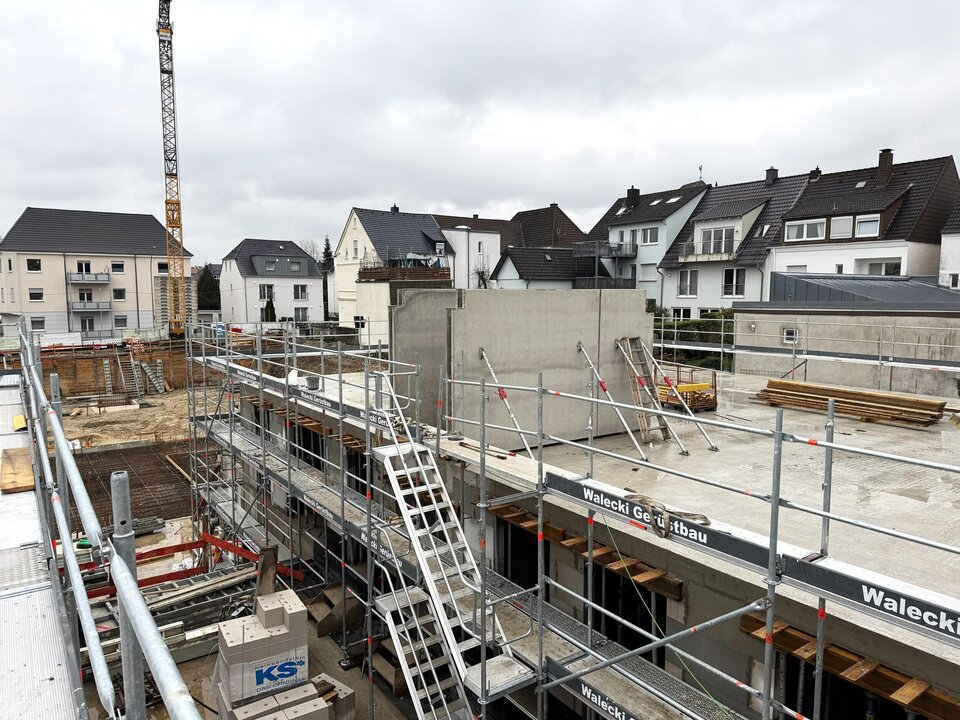 Die belebte Baustelle im Paderborner Querweg zeigt ein im Bau befindliches Betongebäude, umgeben von Gerüsten, einem Kran und einer Mischung aus Wohn- und Geschäftshäusern.