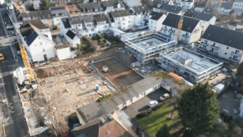Die Luftaufnahme zeigt die belebte Baustelle Querweg mit einer großen Betonplatte, Gerüsten und einem gelben Kran, umgeben von Wohnhäusern und der Straße.