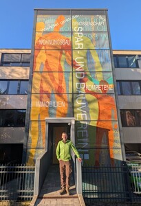 Thomas Milkereit in grüner Jacke steht vor dem modernen Verwaltungsgebäude an der Giersmauer mit einer großen Glasfassade, auf der ein buntes Wandgemälde und die Worte "Wohngsbau" und "Kompetenz" in deutscher Sprache zu sehen sind.